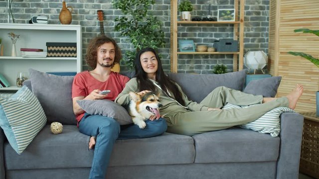 Young family and corgi dog are watching TV on couch at home laughing relaxing enjoying free time together. Entertainment, people and lifestyle concept.