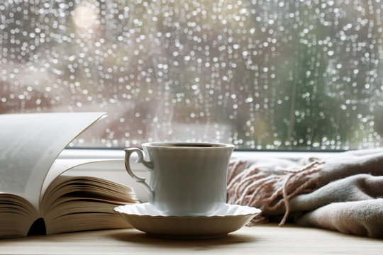 Porcelain cup with hot tea, soft blanket and open book by the window.
