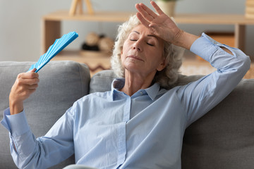 Tired senior woman waving fan feel hot sit on sofa