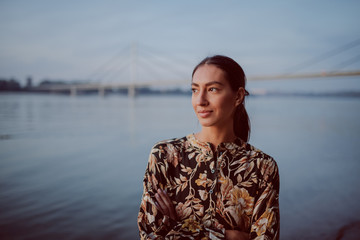 Fototapeta premium Beautiful caucasian brunette in floral dress standing on shore with hands folded and looking away. In background is bridge.