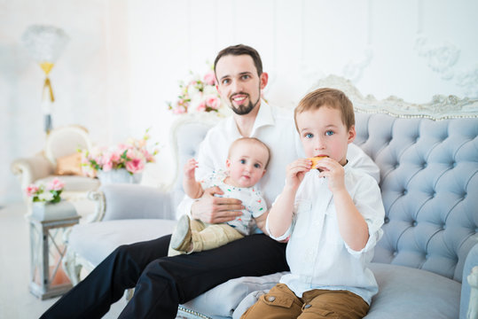 Positive Young Father Is Sitting On The Blue Couch With His Little Sons In A Beautiful Designer Room. The Concept Of Procreation And Family. Father And Son Concept