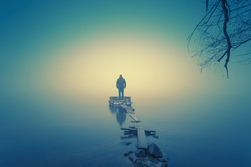Lonely man, standing on the wooden jetty in the autumn foggy river with stones gangway. 