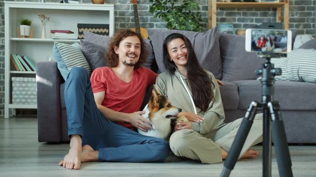 Couple of vloggers Caucasian man and Asian girl are recording video at home caressing corgi dog using smartphone. Lifestyle, pets and vlogging concept.