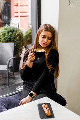 beautiful girl in black sweater drinks cappuccino in cafe by the window