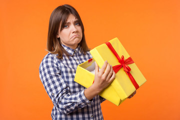 Portrait of unhappy young woman with brown hair in casual checkered shirt holding gift box, looking...