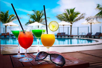 Четыре бокала с разноцветными коктейлями стоят на столе. Рядом лежат солнцезащитные очки. вся композиция на тропическом фоне. Бассейн, пальмы и лежаки.