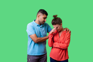 Portrait of unhappy worried young couple in casual wear standing together, man comforting crying woman, holding her shoulders, expressing sympathies. isolated on green background, indoor studio shot