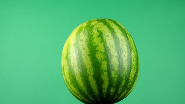 Lit from either side, a large, ripe watermelon rotates slowly in front of a bright green background