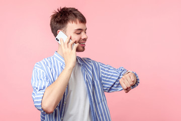 I'll be late! Portrait of careless brown-haired man with small beard in casual shirt talking on phone and looking time on his wristwatches, saying to be late for meeting, indoor. isolated on pink