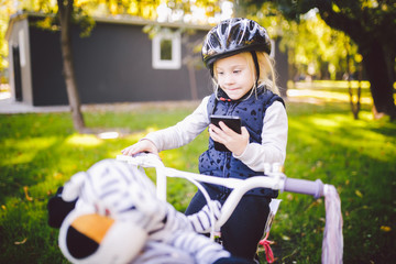 Obraz premium Funny child Caucasian girl blonde in a bicycle helmet near a purple bike with a basket in outside the park on a green lawn grass cart at home. Plays uses hand phone with toothy smile