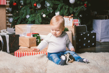Naklejka premium theme winter and Christmas holidays. Child boy Caucasian blond 1 year old sitting home floor near Christmas tree with New Year decor on shaggy carpet skin receives gifts, opens gift boxes in evening