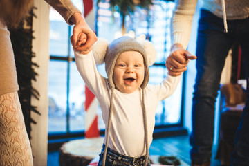 Funny little baby boy 1 year old learning walk home in winter in a decorated New Year house. Young family dad and mom hold by the hands of his son in the loft interior wooden floor near the window