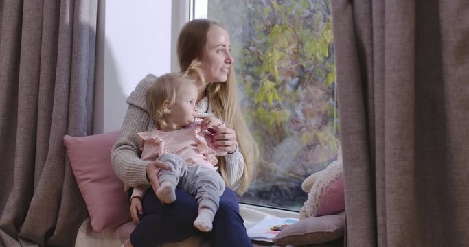 Young beautiful Caucasian mother and daughter sitting at the windowsill and looking out through the window. Woman talking to her child and showing something outdoors. Cinema 4k footage ProRes HQ.