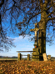 Cross by the road for travellers with a kneeling bench and a large tree in the background in the Czech Republic