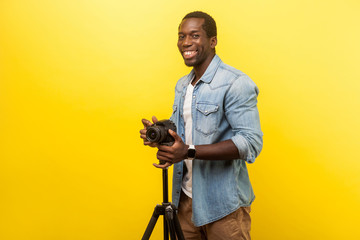 Portrait of excited motivated photographer or traveler in denim casual shirt smiling and posing at camera with professional digital dslr camera on tripod. studio shot isolated on yellow background - obrazy, fototapety, plakaty