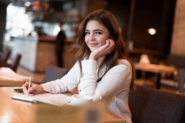 Fototapeta na wymiar young beautiful smiling woman in white jacket taking notes to her planner working outside office drinking hot coffee in the trendy cafe multitasking modern businesswoman