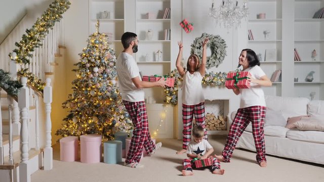 A cheerful European family of four in pajamas is dancing funny with presents in the living room of their home, decorated for Christmas, near the Christmas tree and stairs. A little boy sits on the