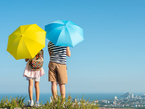 Back Of Boy And Girl Holding Yellow And Blue Umbrella With Top Of City View Background.