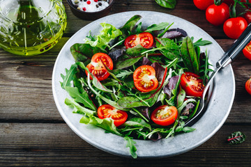 Mix of fresh green salad leaves with arugula, lettuce, spinach, beets and tomatoes on wooden rustic background.