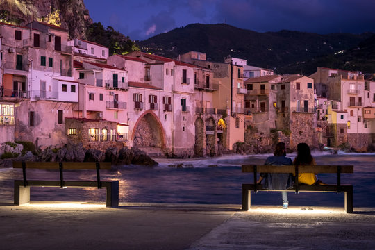 View on habour and old houses in Cefalu at night, Sicily. couple of people resting on a bench