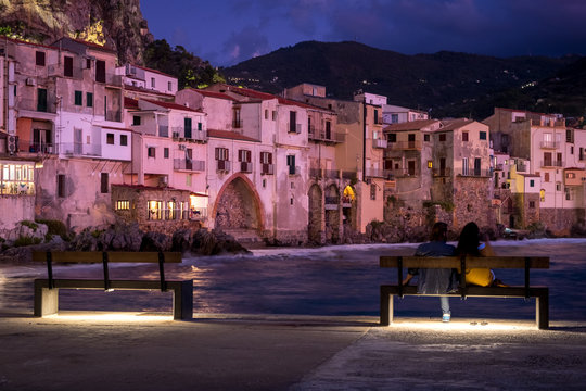 View on habour and old houses in Cefalu at night, Sicily. couple of people resting on a bench
