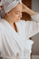 Fototapeta na wymiar Close up of attractive young model in whire bathrobe and soft towel wrapped on head, looking away with satisfied and pleasant expression