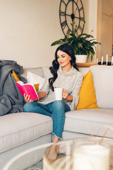Young beautiful woman resting on a sofa, holding cup of tea or coffee, reading a book