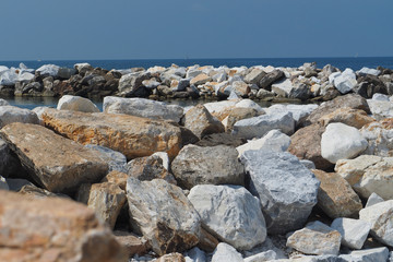 Fototapeta na wymiar White, beige and gray stones against the blue sea with sailboats. Italy, Marina de Pisa