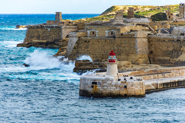 The lighthouse on the Ricasoli Breakwater. In the background waves hit the walls of the Fort...