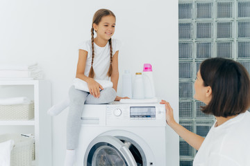 Adorable small girl with pigtails, poses on top of washer, holds white soft towel, looks gladfully at mother, talk about plans after washing. Brunette housewife loads washing machine, being busy