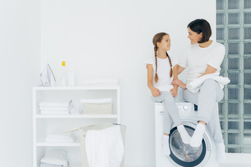 Caring mother talks with daughter, pose on washing machine, surrounded with white linen, do washing...
