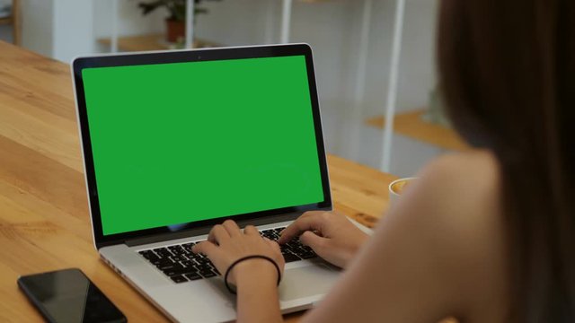 Over the shoulder shot of a business woman working in office interior on pc on desk, looking at green screen. Office person using laptop computer with laptop green screen, sitting at wooden table