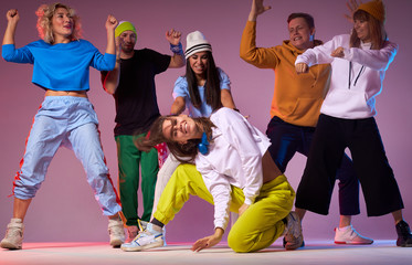 Cheerful sportive street dancers dancing around young wavy haired woman sitting on floor in...