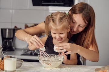 Mother and daughter preparing a sweet cake with flour, milk, sitting on chairs at a table in a...