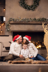 Picture of man and woman in Santa's cap with son sitting on floor on background of fireplace