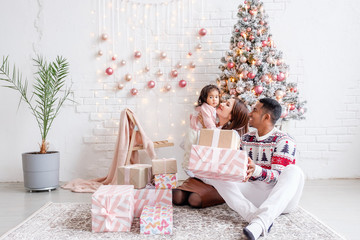 Obraz na płótnie Canvas Happy mixed race family african american dad mom and daughter rejoice celebrating Christmas sitting on living room floor in beautiful bright interior with Christmas tree and decorations. Copyspace
