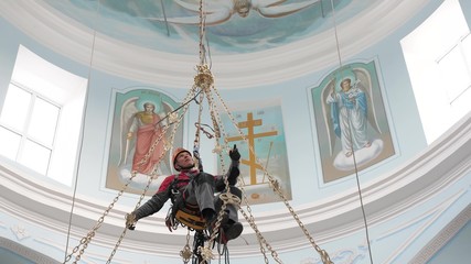 Industrial climber in equipment for high-altitude work working at high altitude in church building. Climber high-altitude installation of a chandelier and lighting. on the climber s belt carabiners. - obrazy, fototapety, plakaty