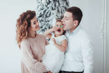 Obraz na płótnie Canvas Happy Family Celebrate New Year Winter Holiday. Young Beautiful Mother Holding Little Daughter on Christmas Tree Background. Pretty Girl with Candy. Funny Caucasian Father Trying to Lick Lollipop