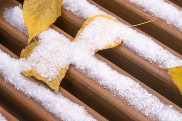 Fototapeta na wymiar Wooden bench seat closeup. Covered with snow and yellow autumn leaves. Selective focus..