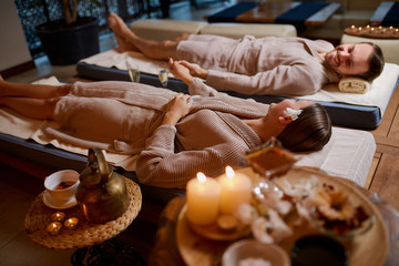 Two young people, lovely couple relax in spa salon together, wearing bathrobes and lying down on desks, after drinking green tea, around candles