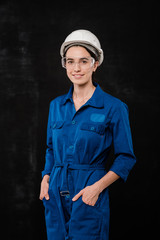 Young female builder in hardhat and blue coveralls keeping hands in pockets