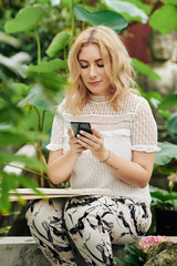 Attractive young blond woman resting in park with sketchbook on her laps and checking smartphone