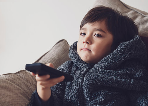 Cute Kid Sitting On Sofa Holding Remote Control, A Happy Child Boy Relaxing At Home Watching TV During Cold Weather Outside In Autumn Or Winter. Indoors Activities For Kids During Bad Weather