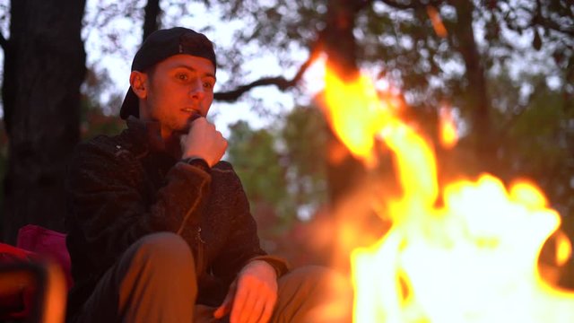 Young handsome man sitting by the campfire in the evening talking to friends (off screen) - focus pull from model to fire