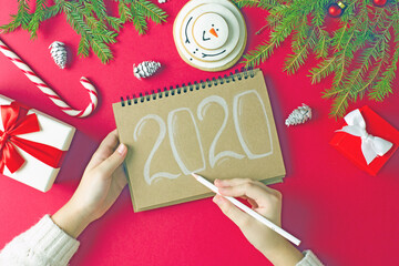 Female hands write with a white pencil the numbers for New Year 2020 on a gray sheet of paper on a red table, next to cones, fir branches, Christmas toys. Christmas background