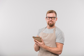 Happy young bearded waiter writing down order of client in notepad