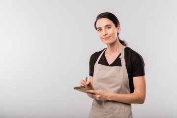 Pretty young brunette waitress in apron making notes about order in notepad