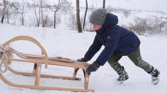 Boy in warm clothing run pushing vintage wooden sled on up the hill on snowy winter day. Little guy sledding in park on snowfall. Tracking shot of child in navy jacket sledge in forest. Slow motion.