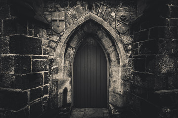 Scary pointy wooden door in an old and wet stone wall building with cross, skull and bones at both sides in black and white. Concept mystery, death and danger. - Powered by Adobe
