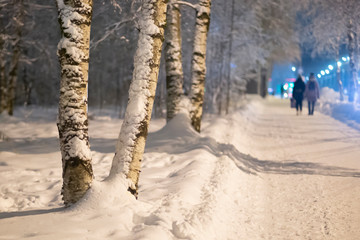 People go at night in winter through the city on a snowy road.